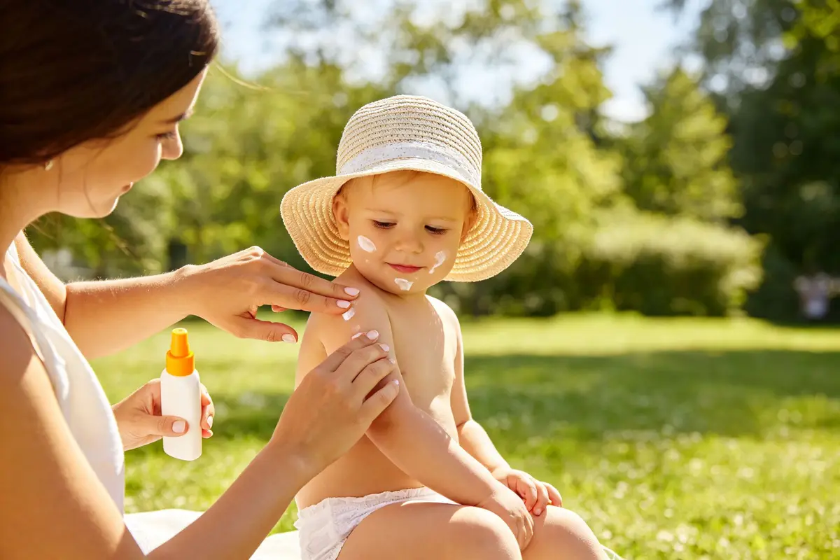Mother applying sunscreen to baby's arms outdoors with sun hat
