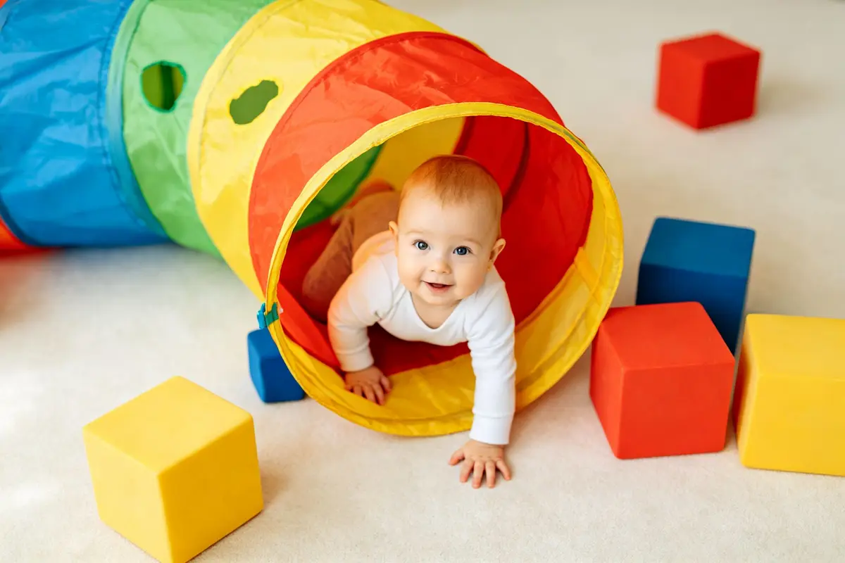 Happy baby crawling through a colorful fabric play tunnel with soft blocks around on a clean carpet