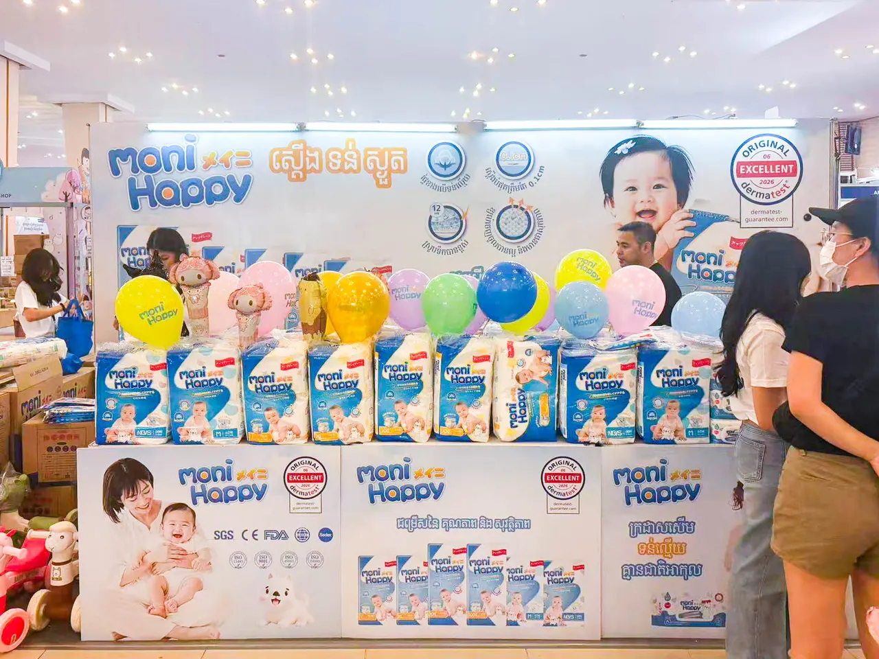 Moni Happy brand booth at the Cambodia Baby & Maternity Expo in Phnom Penh, with the full product lineup displayed and Dermatest® “Excellent” certification featured on the backdrop.