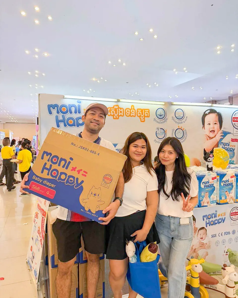 A Cambodian couple posing with a Moni Happy baby diaper pack at the expo booth, holding balloons and smiling with the brand displays behind them.