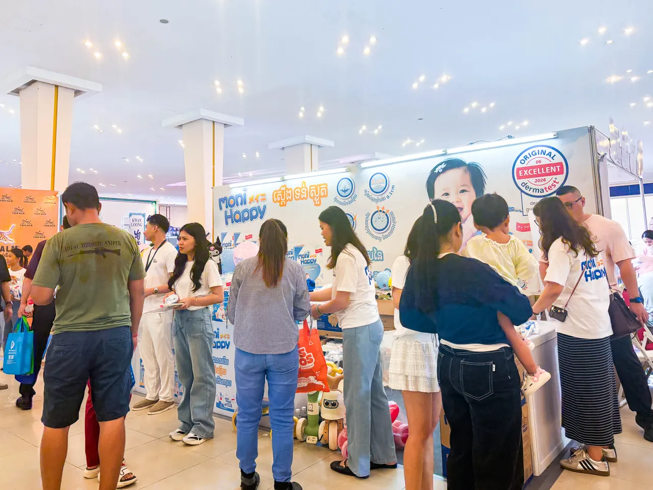 Moni Happy booth staff in branded T-shirts interacting with a Cambodian family during the product trial area of the Cambodia Baby & Maternity Expo.