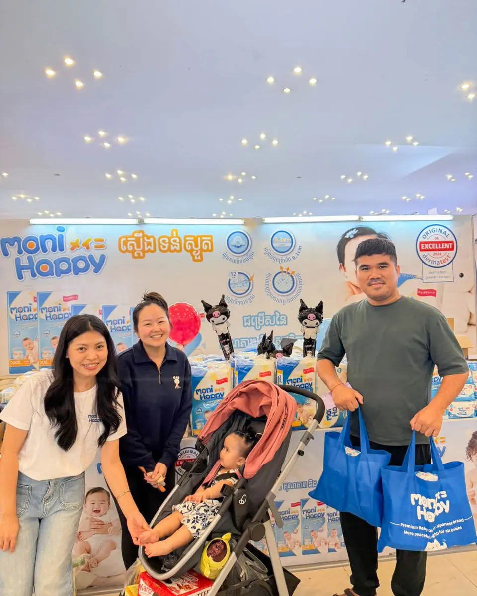 A young Cambodian family at the Moni Happy booth — a mother pushing a stroller with their toddler while the father carries two Moni Happy shopping bags in front of the product display.