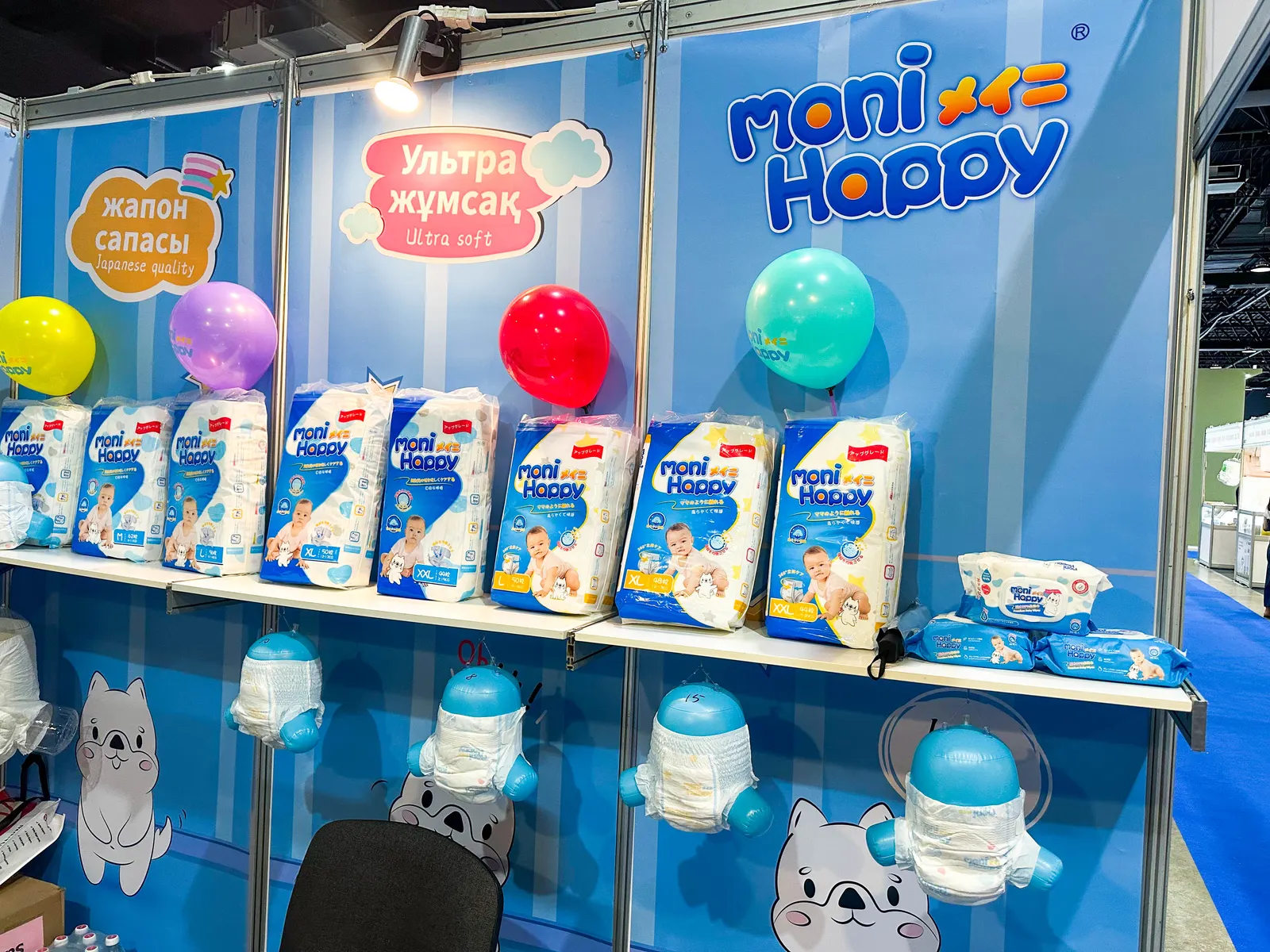 Moni Happy booth at the Kazakhstan Baby & Kids Expo — the full star product lineup displayed on bright blue shelving with the brand mascot and Kazakh-language signage.